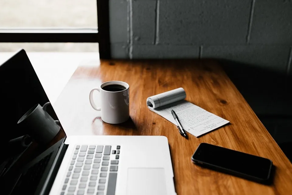 A phone face down on a wooden desk beside a notepad and a cup of coffee